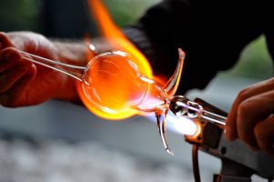 Glass artist produces souvenir objects by melting glass with heat.